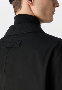 Veste noire avec un col haut, tissu texturé et une étiquette de marque. Le design comprend des coutures détaillées et une silhouette structurée.