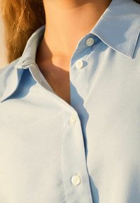 Camicia azzurra chiara con colletto in tessuto morbido, dotata di una chiusura a bottoni frontale e due bottoni bianchi, che mette in evidenza un design pulito e semplice.