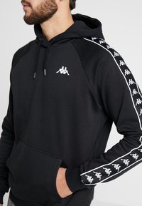 Sudadera negra con bolsillo canguro, logo blanco en el pecho y rayas blancas en contraste con un logo repetido a lo largo de las mangas.