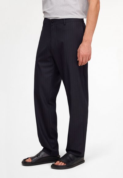 Homme portant un pantalon de costume noir à fines rayures et des sandales noires à bout ouvert, debout devant un fond blanc uni.