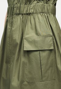 Υφασμα σε ελιάς πράσινο με ελαστική μέση και μεγάλη ορθογώνια μπροστινή τσέπη με κλείσιμο καπακιού.