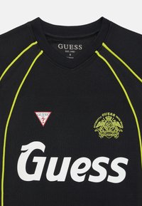 Svart sportsskjorte med gule detaljer, v-hals og en stor hvit "Guess" Logo. Inkluderer røde og gule merker på forsiden.