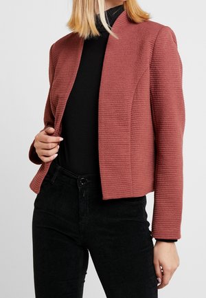 Vrouw draagt een gestructureerde stoffige rozenblazer over een zwarte coltrui en zwarte broek, houdt de blazer met één hand open.