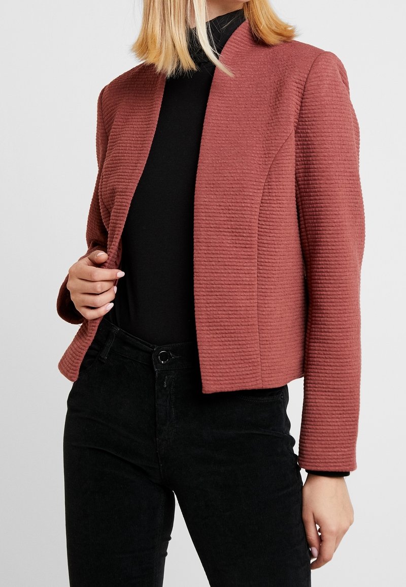 Femme portant un blazer rose poudré texturé sur un col roulé noir et un pantalon noir, tenant le blazer ouvert d'une main.