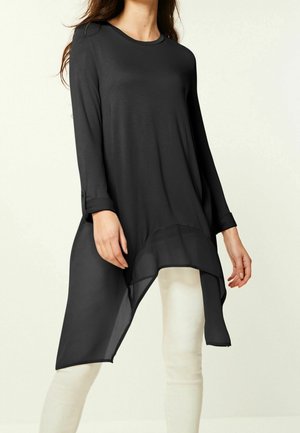 Femme portant une longue tunique noire asymétrique à manches longues sur un pantalon blanc, sur un fond clair uni.