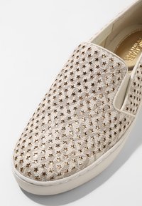 Chaussure à enfiler en tissu beige avec des découpes en forme d'étoiles dorées, une surface texturée, et une semelle en caoutchouc blanche. L'intérieur présente un logo doré.