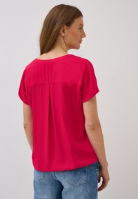 Blouse fuchsia avec des manches courtes retroussées, détails de plis au dos et texture lisse, associée à un jean bleu clair.