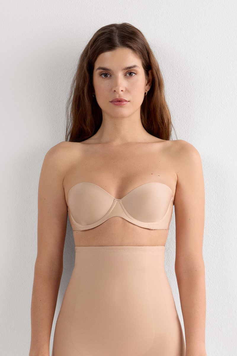 Vrouw met lang bruin haar die een strapless huidkleurige beha en een high waist huidkleurige corrigerende onderkleding draagt tegen een effen witte muur achtergrond.