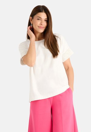 Woman with long brown hair wearing hoop earrings, a white short-sleeve top, and bright pink wide-leg pants against a plain background.