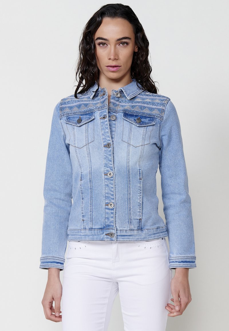 Világoskék farmerdzseki, testhezálló fazonnal, gombos elülső rész, két mellzseb és bonyolult varrás a vállon.