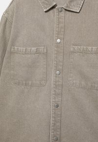 Bež denim srajca z dvema prsnikoma žepoma in kovinskimi pritiskači spredaj, s klasičnim ovratnikom in vidnim šivom.