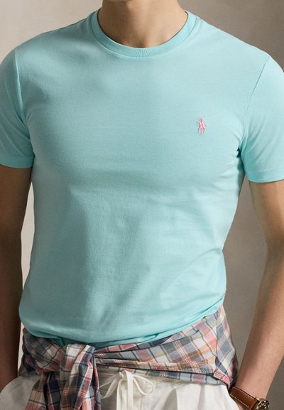 Άνδρας που φοράει ανοιχτό μπλε μπλουζάκι με στρογγυλή λαιμόκοψη και ροζ κεντημένο λογότυπο, πουκάμισο καρό δεμένο γύρω από τη μέση, λευκό παντελόνι και καφέ ρολόι χειρός.