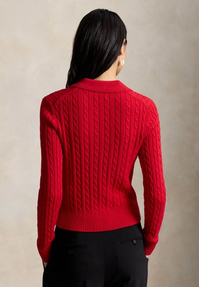 Kobieta o długich ciemnych włosach, ubrana w czerwony sweter z grubym splotem i czarne spodnie, stojąca tyłem z rękami w kieszeniach na beżowym tle.