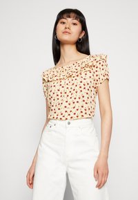 Blusa floral com design de ombros à mostra e babados, apresentando um fundo amarelo claro com padrões de flores vermelhas e verdes. Usada com calças brancas.