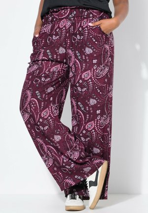 Person iført løstsiddende bordeaux bukser med paisley-mønster og sidelommer samt hvide sneakers med sorte detaljer, stående op ad en ensfarvet lys væg.