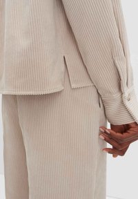 Camicia in corduroy beige con righe verticali, polsini con bottoni e spacchi laterali; abbinata a pantaloni coordinati, che mostrano una texture morbida e toni neutri.
