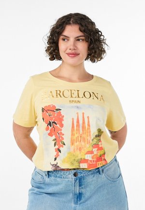 Frau mit lockigem Haar, die ein gelbes Barcelona Spanien T-Shirt mit einer bunten Stadtszenerie und Blumen trägt, kombiniert mit blauen Jeans.