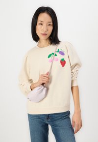 Beige pulóver hímezett gyümölcs mintákkal, kerek nyakkivágással és puffos ujjakkal. Egy kis levendula táskával és kék farmernadrággal párosítva.