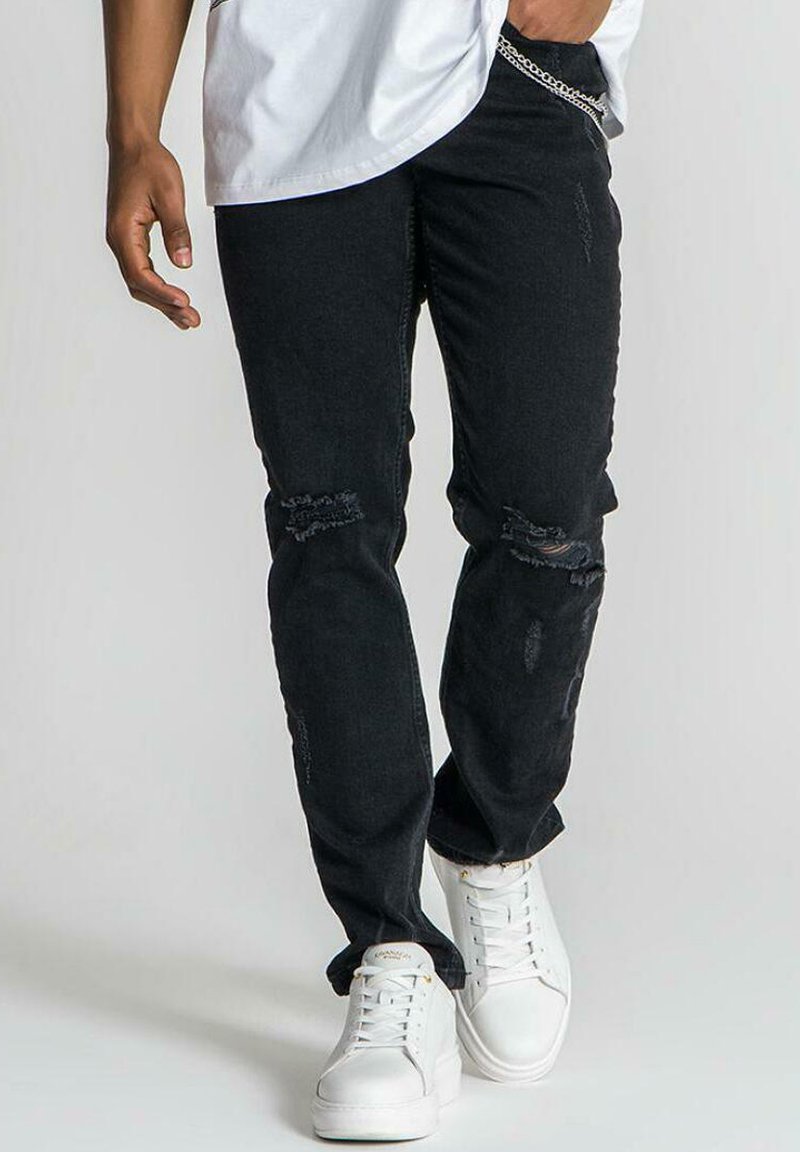 Jeans negros desgastados con rotos, corte recto, acompañados de zapatillas blancas. El look presenta un diseño casual y un detalle minimalista en cadena de plata.