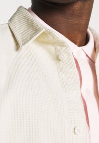 Primer plano de una persona que lleva una camisa abotonada texturizada color crema sobre una camisa de cuello color rosa claro.