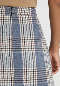 Jupe à carreaux bleu, blanc et marron taille haute portée avec un haut beige, montrant une fermeture éclair latérale et une main reposant près de la hanche.