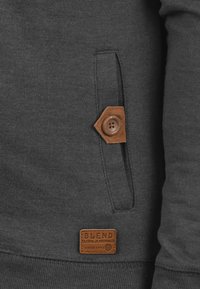 Dark gray sweatshirt featuring a leather patch with text, a button detail near a side pocket, and a textured fabric finish.