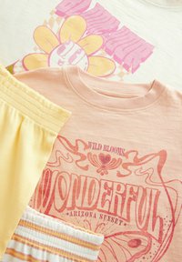Drie kledingstukken plat gelegd: een crèmekleurig shirt met een bloemenafdruk, een roze shirt met retro tekst, en gele gestreepte shorts met een elastische tailleband.