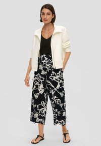Veste blanche zippée, débardeur noir à côtes, pantalon large noir à motif floral blanc, et sandales plates noires. Fond neutre.