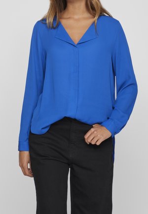 Femme portant un chemisier bleu vif à manches longues avec un col en V et un pantalon noir, debout, ajustant l'ourlet de la chemise d'une main.