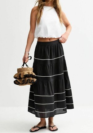 Mujer con top corto sin mangas blanco, falda midi negra con rayas blancas, chanclas negras, sosteniendo un bolso tejido con volantes negros.