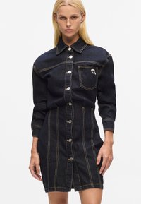 Dunkelblaues Denim-Hemd Kleid mit langen Ärmeln, knöpfbarem Frontverschluss, Brusttasche und kontrastierendem gelben Nähten. Hat eine taillierte Silhouette und endet auf Kniehöhe.