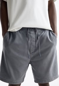 Szare bawełniane szorty z elastycznym pasem, regulowanym sznurkiem, bocznymi kieszeniami i gładką fakturą. Noszone z białą koszulką.