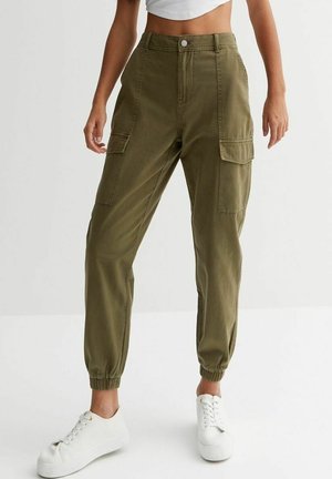 Femme portant un pantalon cargo vert olive avec des chevilles élastiques, des poches à rabat latérales, des baskets blanches et un haut blanc court sur fond uni.