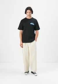 Sort bomulds T-shirt med et blåt grafisk print med teksten "IRON MAIDEN" på brystet, parret med cremafarvede vidde bukser og sorte sneakers.
