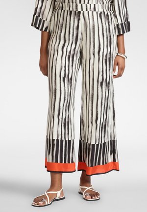 Donna con pantaloni a gamba larga a righe verticali bianco e nero con bordo arancione, sandali bianchi con cinturini e bracciale argentato al polso destro.