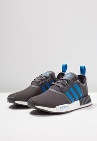 Grå mesh-sportskor med blå ränder, vit sula och en texturerad yta. Har en dragflik på hälen och snörningsdesign.