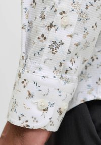 Lähellä näkyvä valkoinen, teksturoitu pitkähihaisen paidan hihansuu, jossa on pieniä ruskeita ja sinisiä kukkakuoseja sekä valkoisia nappeja, tummapintaisella kädellä.