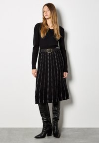 Suéter negro de canalé con mangas largas, combinado con una falda midi negra con estampado y un cinturón ancho con hebilla dorada. Botas negras hasta la rodilla.