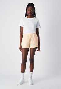Weißes Baumwoll-T-Shirt mit kurzen Ärmeln und pinkem "Champion"-Logo. Helle Pfirsich Drawstring-Shorts. Weiße Sneaker und Socken.
