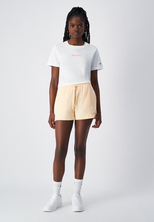 Camiseta de algodón blanca con mangas cortas y logo "Champion" en rosa. Pantalones cortos de tiro medio en color melocotón claro con cordones. Zapatillas y calcetines blancos.