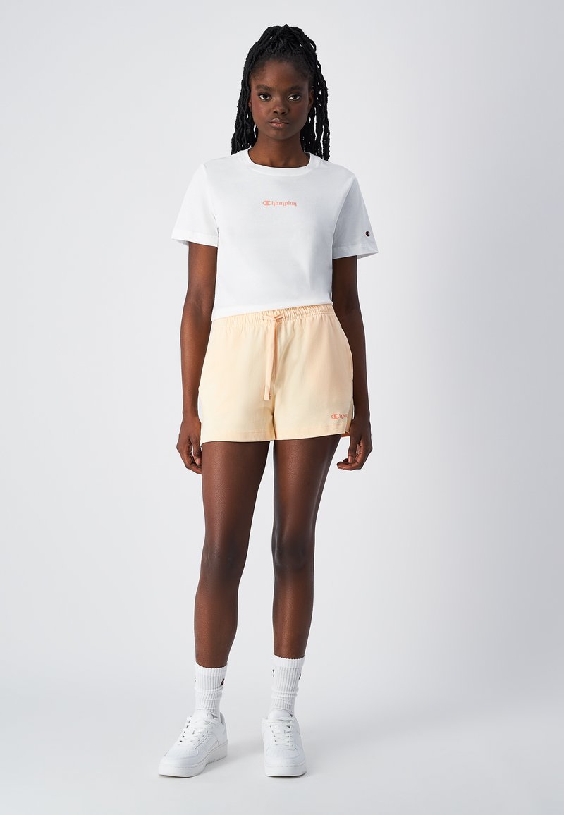 Weißes Baumwoll-T-Shirt mit kurzen Ärmeln und pinkem "Champion"-Logo. Helle Pfirsich Drawstring-Shorts. Weiße Sneaker und Socken.