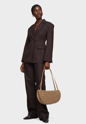 Kobieta ubrana w ciemnobrązową, dopasowaną marynarkę i spodnie, trzymająca beżową torebkę na ramię z fakturą, z dwoma paskami.
