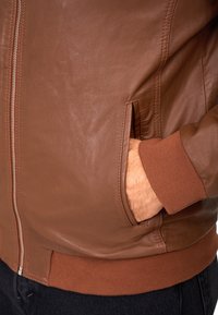 Bruine leren jas met een gladde textuur, geribbelde boorden, een rits aan de voorkant en zijzakken. Heeft contrasterende stiksels.