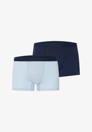 Muskelöser Mann steht barfuß und blickt nach vorne, trägt hellblaue Boxer mit einem dunklen Bund vor einem schlichten hellen Hintergrund.