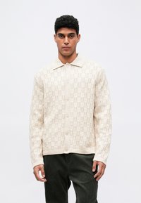 Lys beige strikkegenser med hevet mønster, full knappestolpe og myk krage. Teksturert stoff med ribbede mansjetter og nederkant.