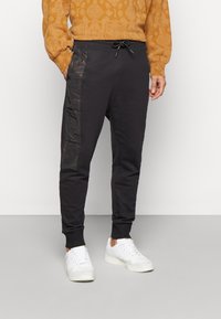 Sorte joggers med en tætsiddende design, der har et skinnende sort sidepanel, elastiske manchetter og en snøre i livet, parret med hvide sneakers.
