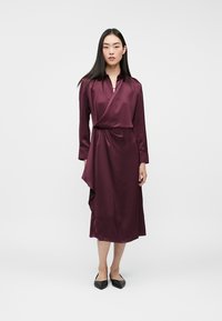 Kobieta w długiej, burgundowej satynowej sukience w stylu kimono oraz czarnych czółenkach na szpilce, stojąca na tle jednolitego, jasnoszarego tła.