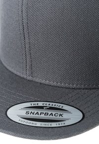 Casquette snapback grise avec un tissu texturé, visière plate et un patch logo rond portant l'inscription "THE CLASSICS SNAPBACK YUPOONG SINCE 1974."