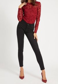 Haut à manches longues en dentelle rouge, jean noir ajusté, chaussures à talons rouges avec un accent volanté. Fond simple ; met en valeur les détails de la tenue.