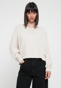 Žena s rovnými tmavými vlasy, oblečená v oversized dlouhém světle béžovém tričku s dlouhým rukávem a černých kalhotách s vysokým pasem, s rukama v kapsách, na neutrálním pozadí.